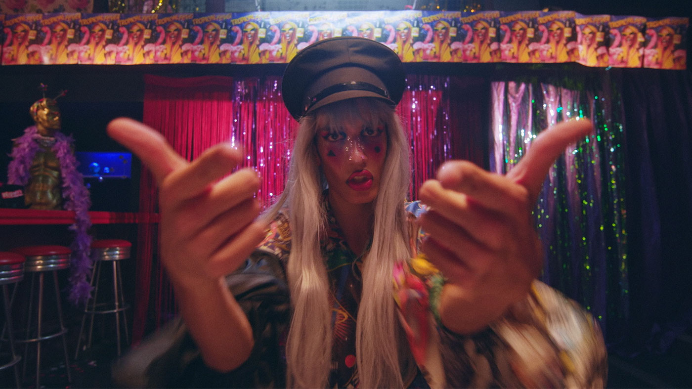 A person wearing a blonde wig and colourful outfit points their fingers towards the viewer; they are in a bar with a sparkly background