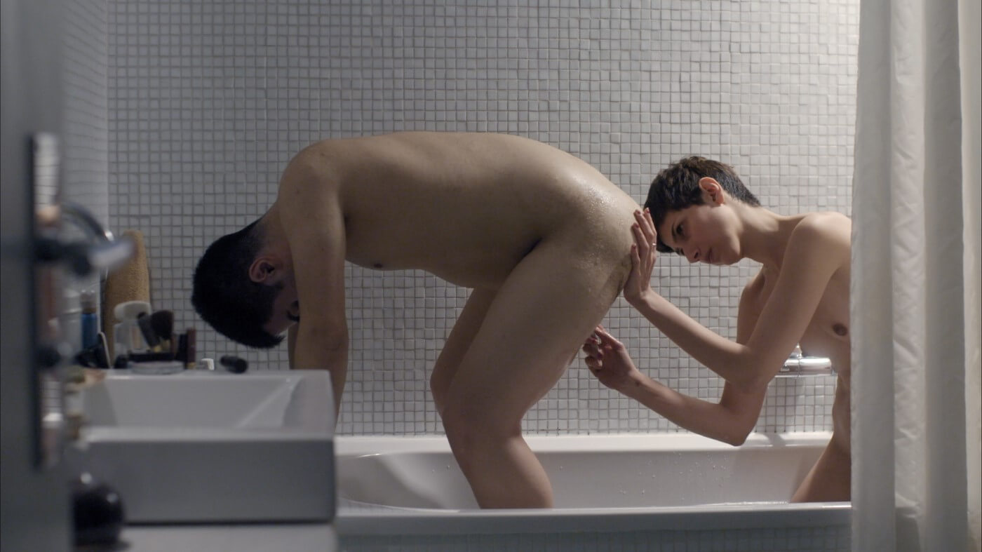 Two naked people crouched in a bathtub, one is bent over and the other shaves them from behind