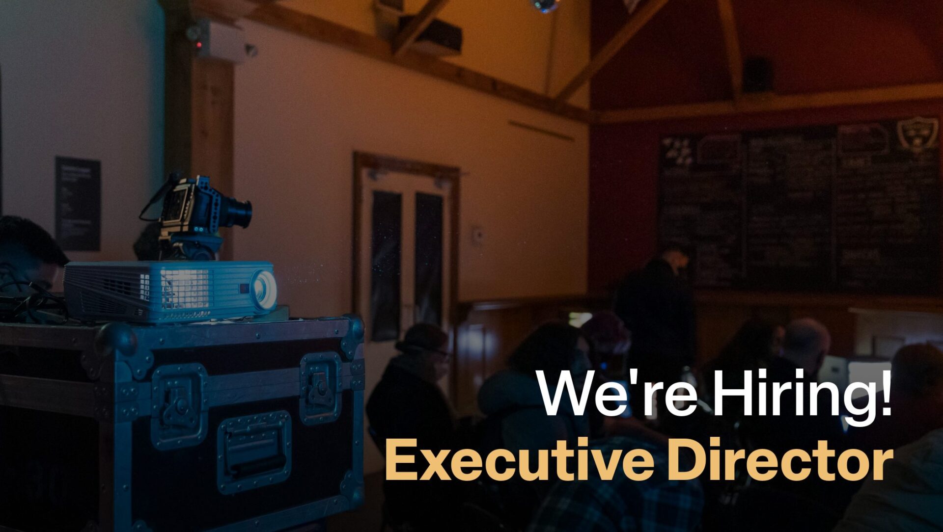 Text reads "We're Hiring: Executive Director" over a photograph of a dimly lit bar with a projector and camera on the left.